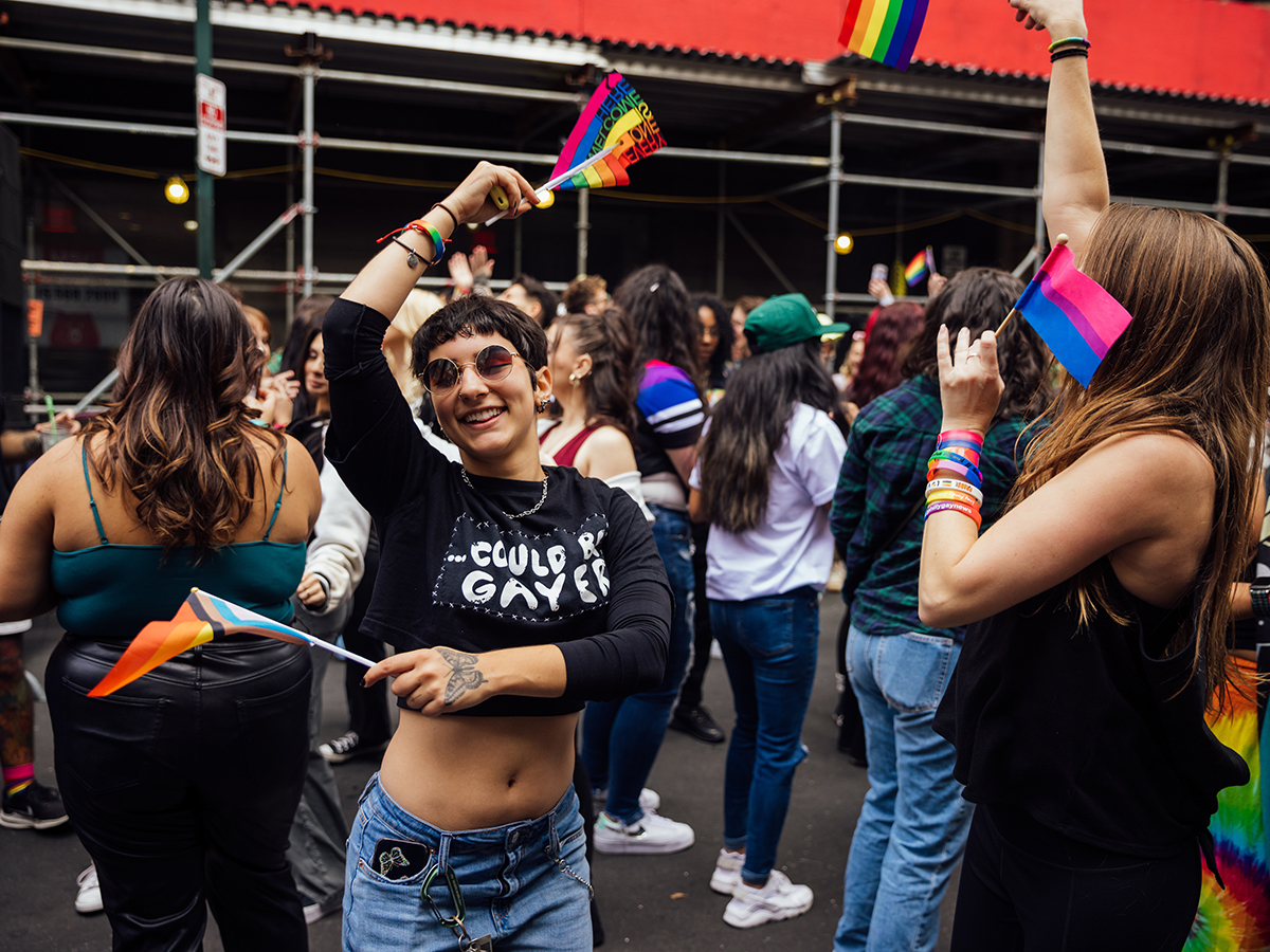 A joyful person waves mini rainbow and trans pride flags in a crowded Philly Pride celebration.